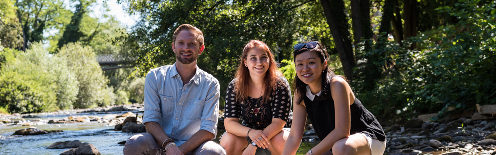 Three students are in the forest next to a small stream. They are squatting on the ground and smiling for a group photo.